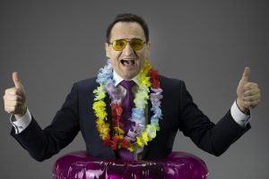 happy businessman dressed in a formal suit and lei Hawaiian welcome necklace in inflatable swim ring gesticulating in sunglasses isolated on gray background . holiday concept